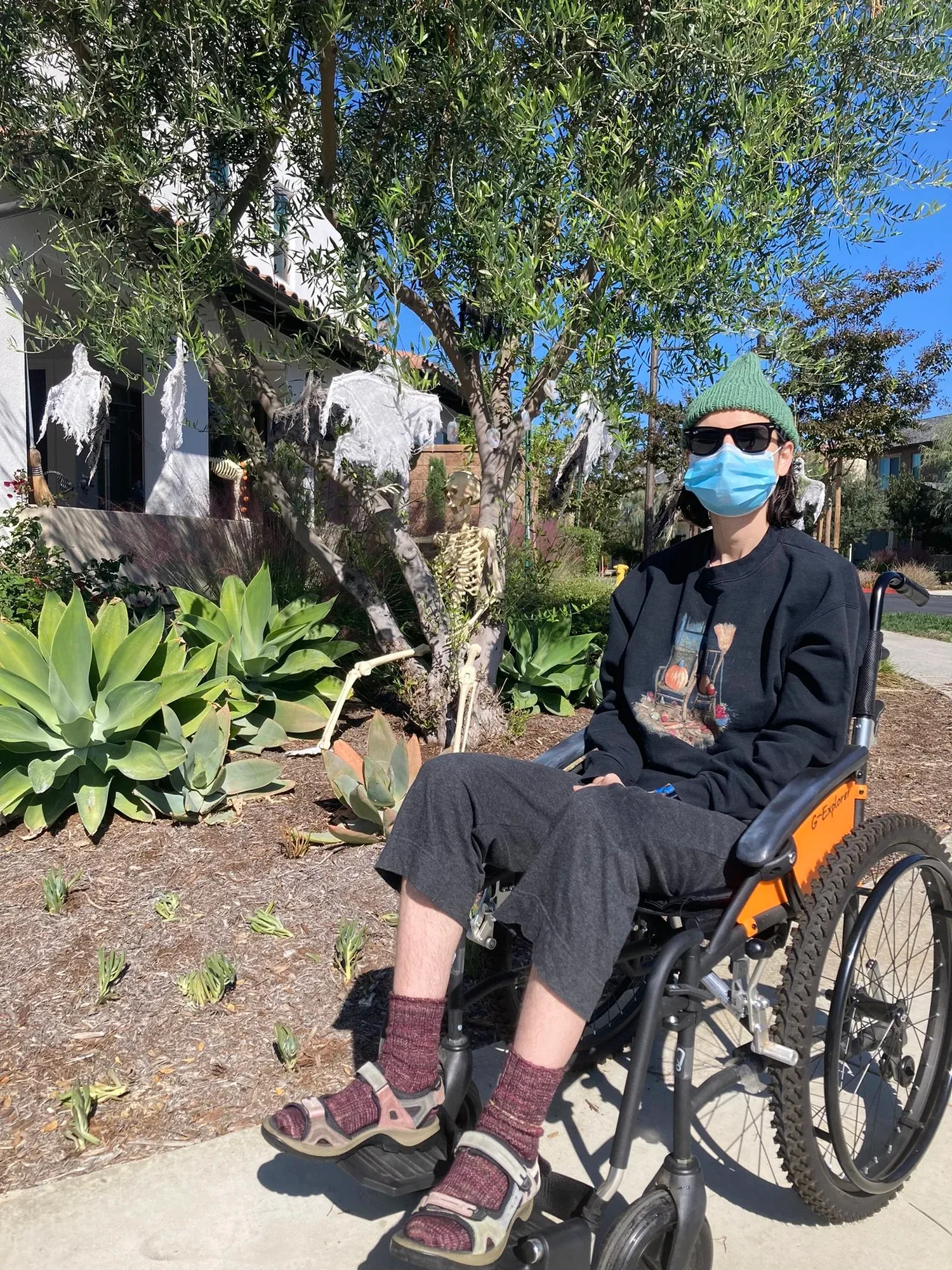 A young person is sitting in a wheelchair outside while wearing a mask.