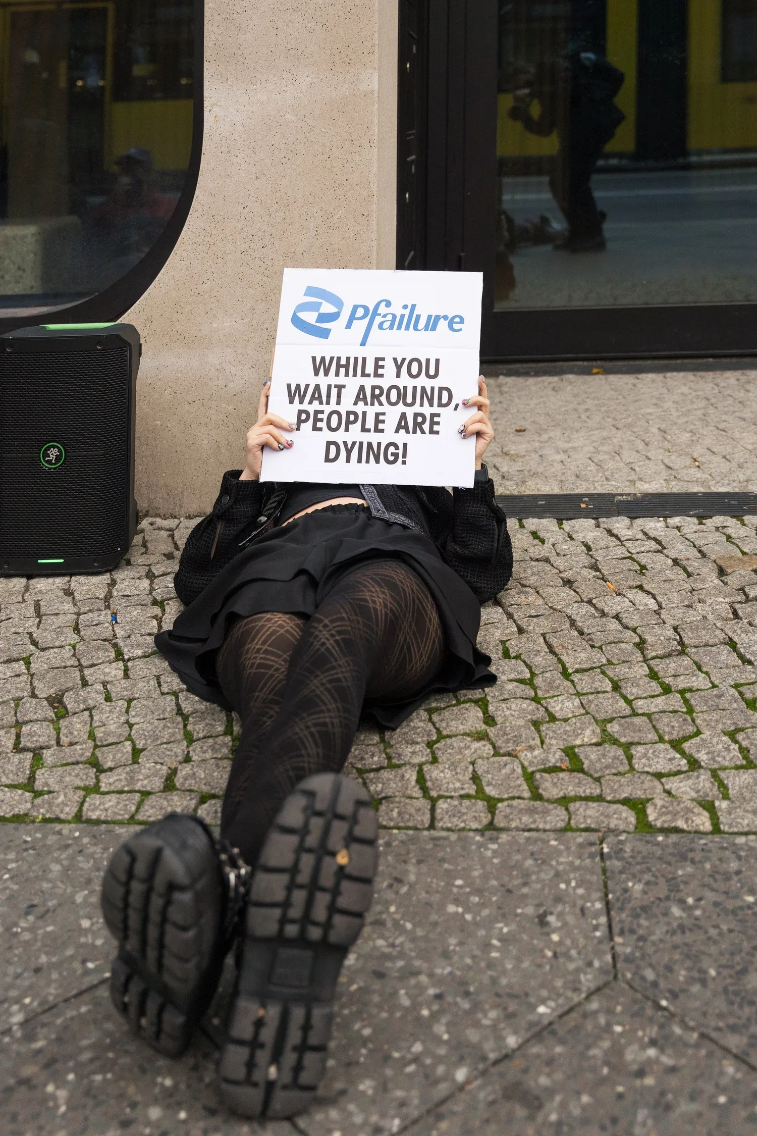 A protester lies on the pavement holding up a sign that reads “Pfailure: While you wait around, people are dying!”