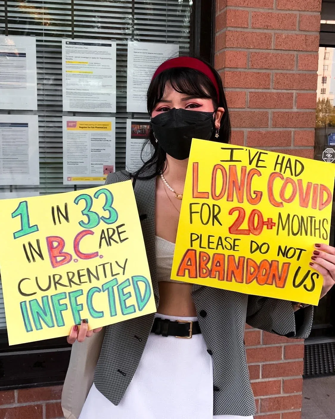 A young woman is wearing a mask outside while holding two protest signs which advocate for covid protections and long covid treatments.