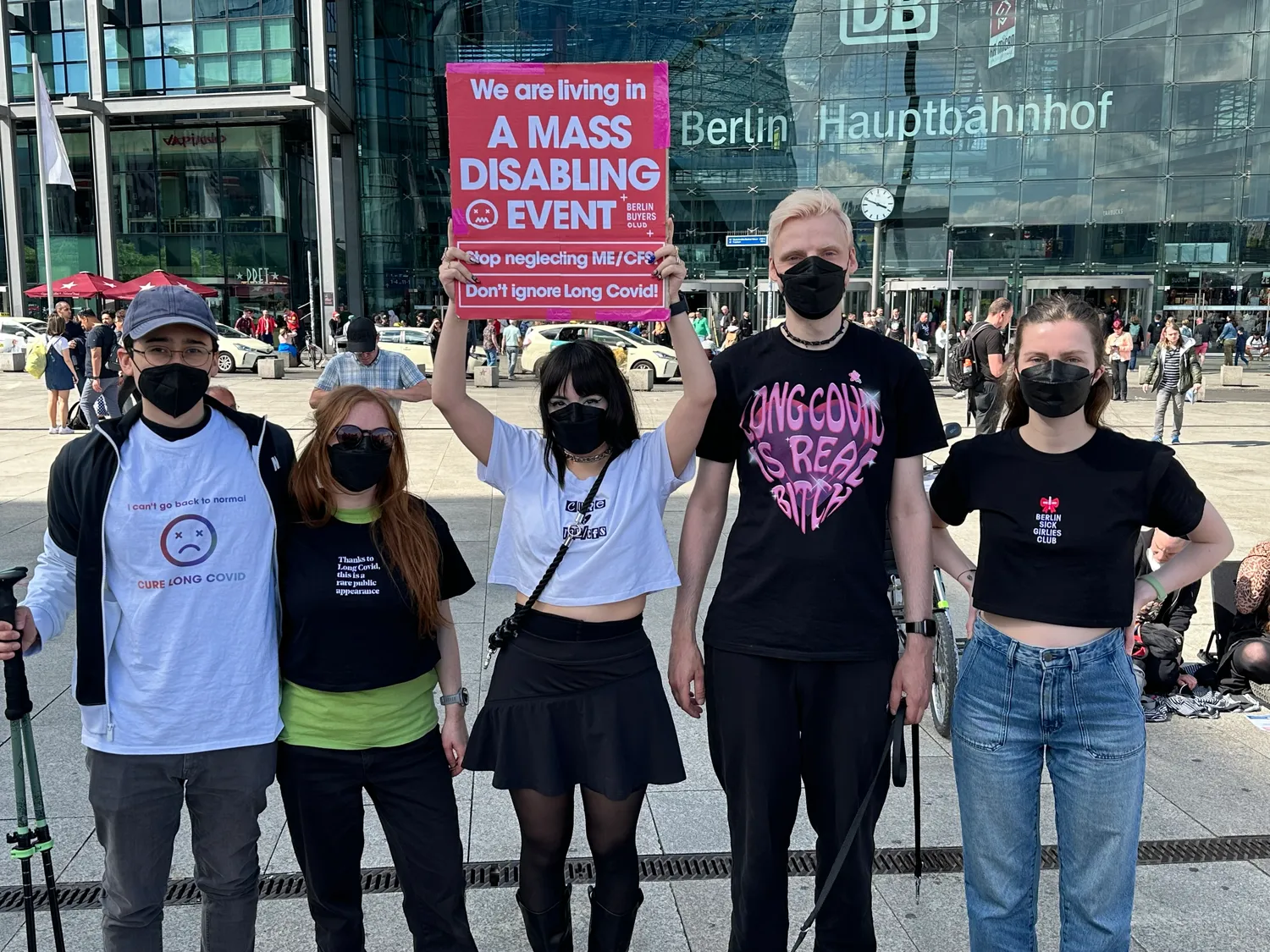 Five masked Berlin Buyers Club members outside Berlin Hauptbahnhof, one holding up a sign: “We are living in a mass disabling event. Stop neglecting ME/CFS. Don’t ignore Long Covid!”