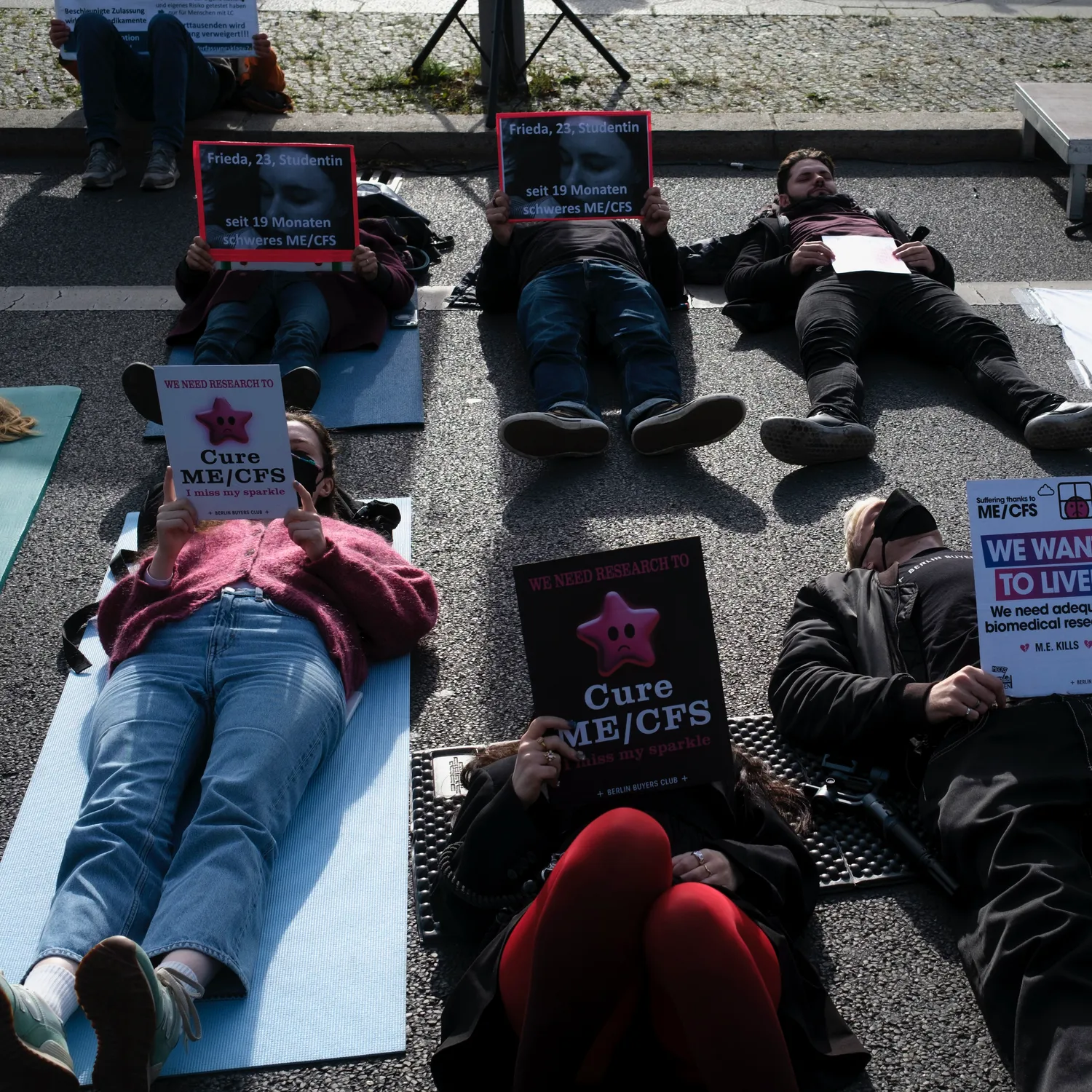 People lying on mats in the street at a Berlin Buyers Club protest, holding signs that read “We need research to cure ME/CFS, I miss my sparkle” and “Frieda, 23, Studentin, seit 19 Monaten schweres ME/CFS”.