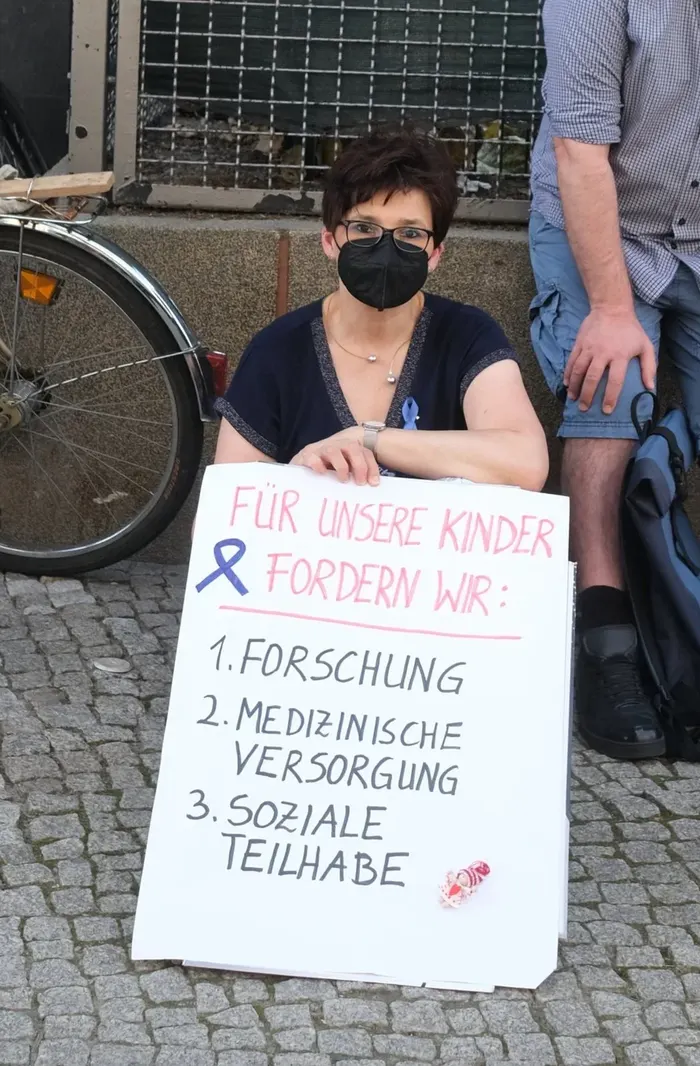 A masked woman sits by a sign: Für unsere Kinder fordern wir Forschung, medizinische Versorgung, soziale Teilhabe