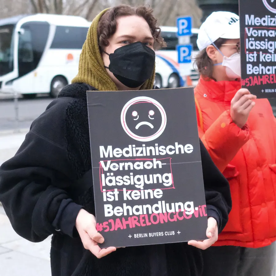 A masked protester holds a sign: Medizinische Vernachlässigung ist keine Behandlung