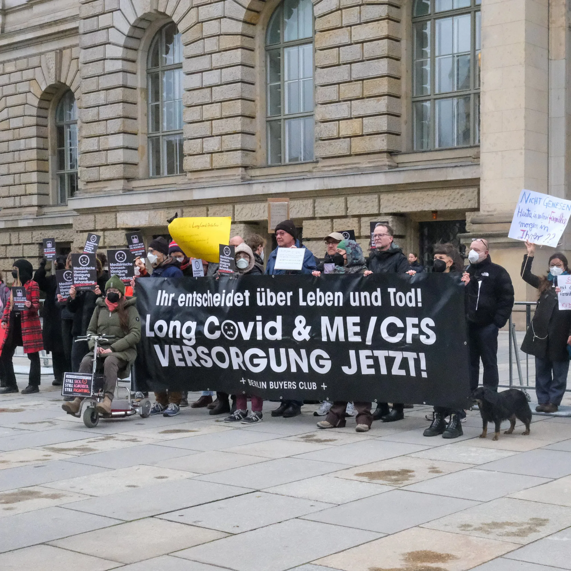Masked protesters outside the Berlin House of Representatives behind a black banner: Ihr entscheidet über Leben und Tod! Long Covid & ME/CFS Versorgung jetzt!