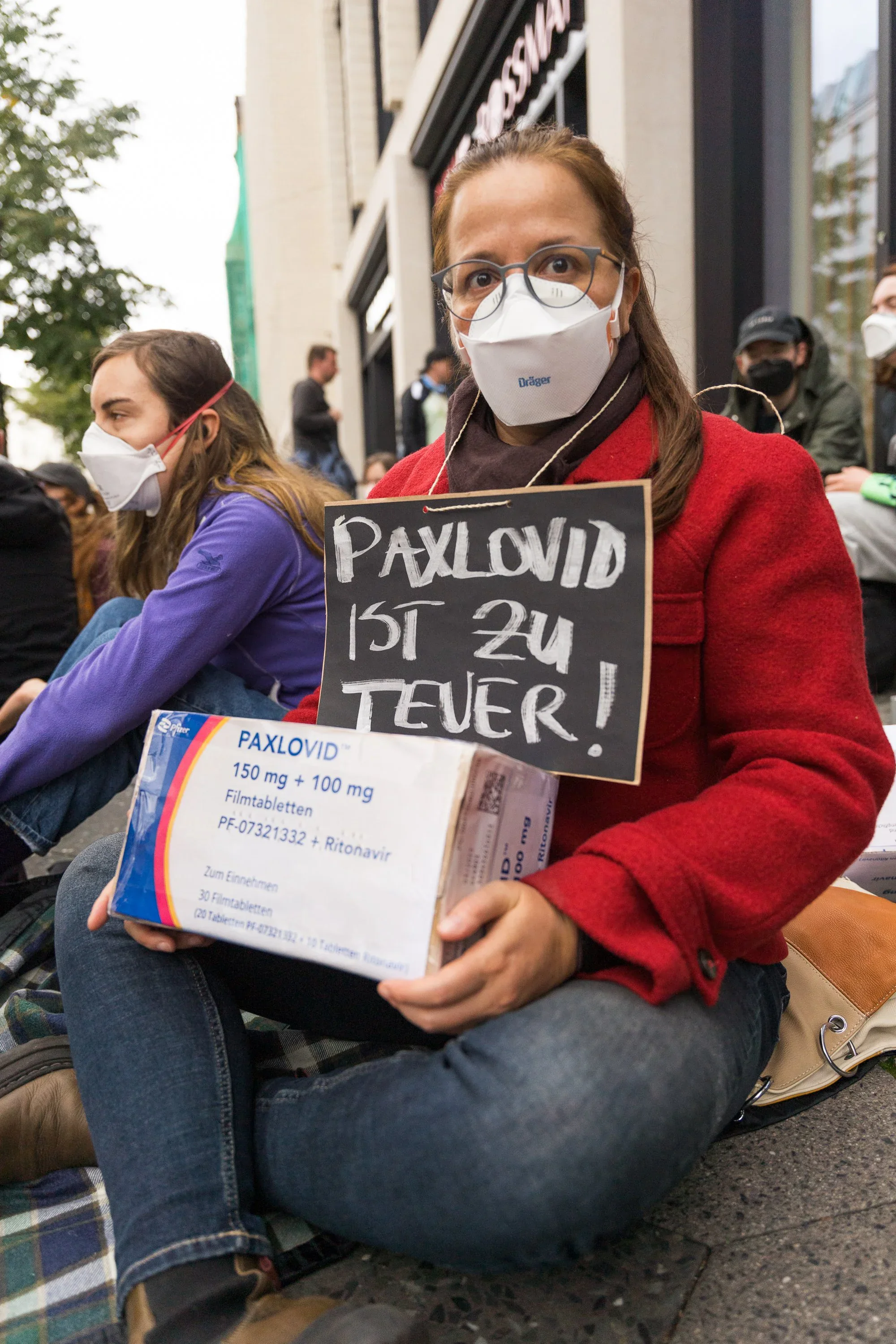 A masked protester sits on the pavement holding a Paxlovid box and a sign reading: Paxlovid ist zu teuer!